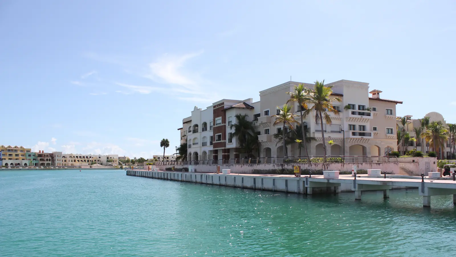 La marina de Cap Cana vista desde el agua: muelle de hormigón y edificios bajos de estuco claro con palmeras entre ellos.