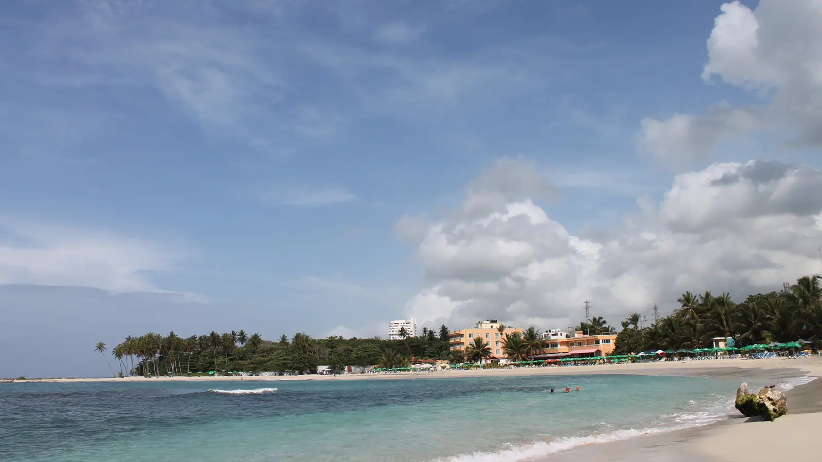 La playa pública de Juan Dolio en una bahía poco profunda, con edificios bajos de colores y sombrillas entre las palmeras al fondo.
