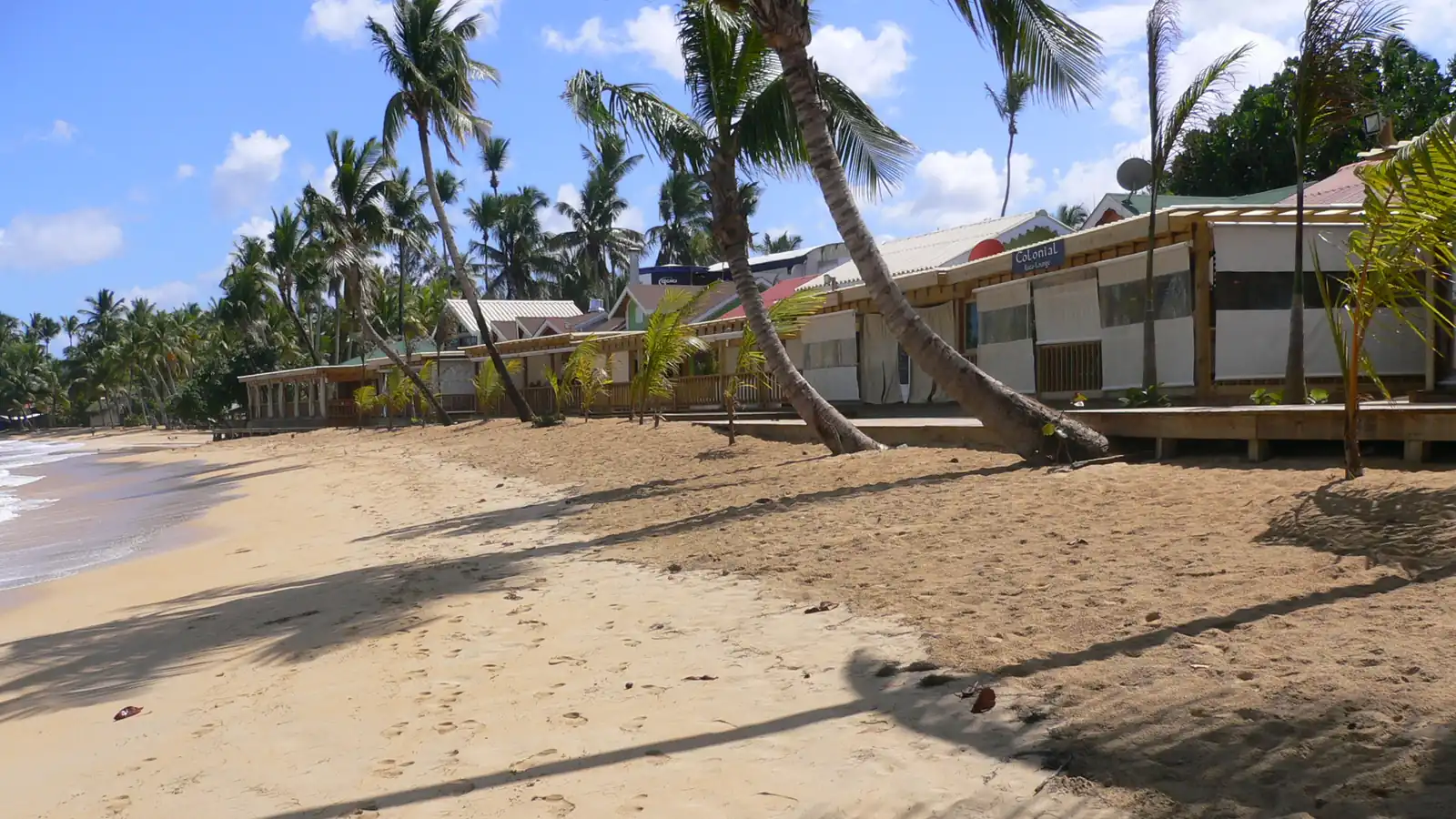 El frente de playa de madera del Pueblo de los Pescadores en Las Terrenas: restaurantes de una sola planta bajo cocoteros inclinados sobre la arena.