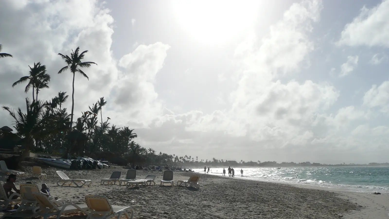 Playa de arena blanca bordeada de palmeras en Punta Cana, con el sol alto entre las nubes y bañistas caminando junto a la orilla.